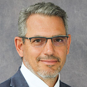 A man with glasses and peppery gray hair stands in front of a gray background.