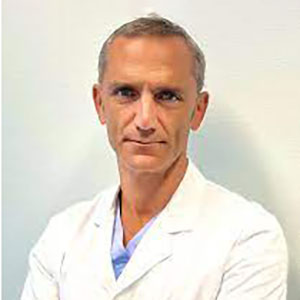 A man in a white doctor's coat stands against a hospital wall.