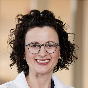 A woman with curly dark hair smiles and wears a white lab coat.