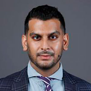 A man with a purple tie and light beard in front of a gray background.